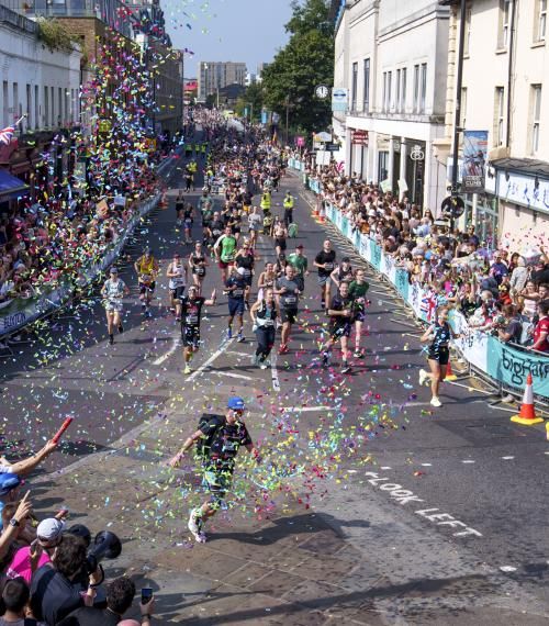 A road is full of people running the Big Half in London. There are spectators standing down either side and someone has fired a confetti cannon